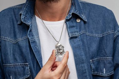 Kot ceketli ve beyaz tişörtlü karizmatik bir adam gri arka planda duruyor. Boynunda şık bir kolye var..