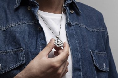 Kot ceketli ve beyaz tişörtlü karizmatik bir adam gri arka planda duruyor. Boynunda şık bir kolye var..