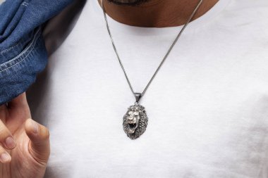 Kot ceketli ve beyaz tişörtlü karizmatik bir adam gri arka planda duruyor. Boynunda şık bir kolye var..