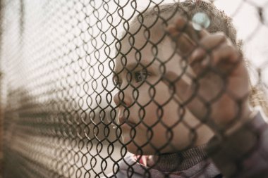 Metal bir çitin arkasında üzgün üzgün bakan küçük bir mülteci kız. Mültecilerin ve yerlerinden edilen insanların sosyal sorunu.