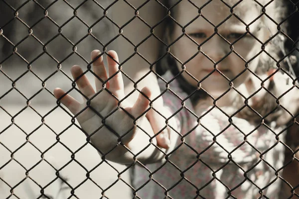 The child's hands are holding onto a metal mesh fence. The problem of forced deportation of Ukrainian children from the territories occupied by Russia.