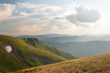 The diverse relief of the Carpathian Mountains with a white spot of snow that has not yet fully melted and a smoky horizon against the background of a gray sky with clouds