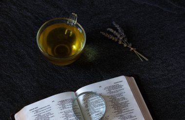 Kutsal Kitap 'ı bir büyüteçle aç, sarı renkli bir fincan sıcak çay, ve koyu granit bir masada kurutulmuş lavanta çiçekleri. Kutsal kitap üzerine Hıristiyanlık. İncil konsepti. Üst görünüm.