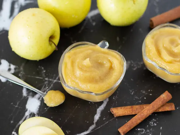 Homemade applesauce (apple puree or mousse) in small glass bowls with organic golden delicious apples and cinnamon sticks on a black textured table. Creamy and healthy baby food.