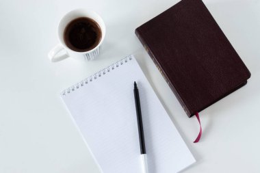 Kapalı bir İncil kitabının olduğu boş bir defter sayfası ve metin için fotokopi alanı olan beyaz arka planda bir fincan kahve. Üst açı görünümü. İncil okuma ve dua etme kavramı..