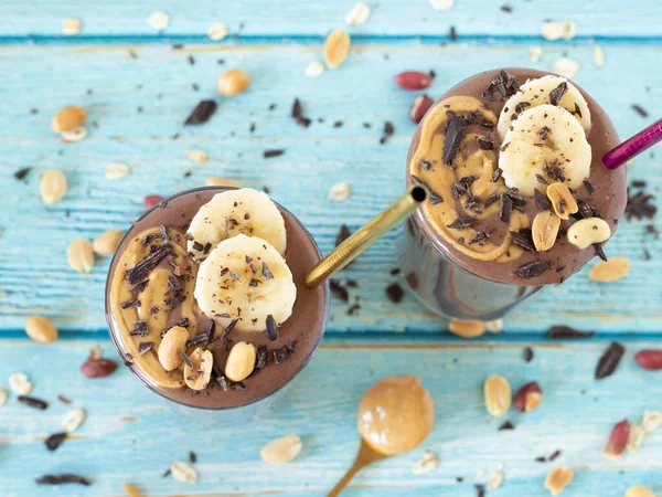 Mavi ahşap arka planda yeniden kullanılabilir pipetlerle iki bardakta çikolatalı fıstık ezmeli muzlu smoothie. Üst görünüm.