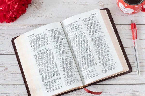 Open Holy Bible Book with fresh red flowers and a cup of a warm coffee cup on a wooden table. Top view, a closeup. The biblical concept of reading and studying the Word of God Jesus Christ.