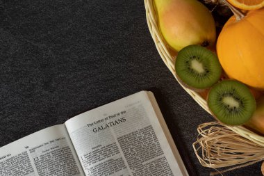 An open Holy Bible book of Galatians with a wicker basket full of various fresh fruits with copy space. The biblical concept of the Christian fruit of the Spirit. Love, joy, peace. Top view. clipart