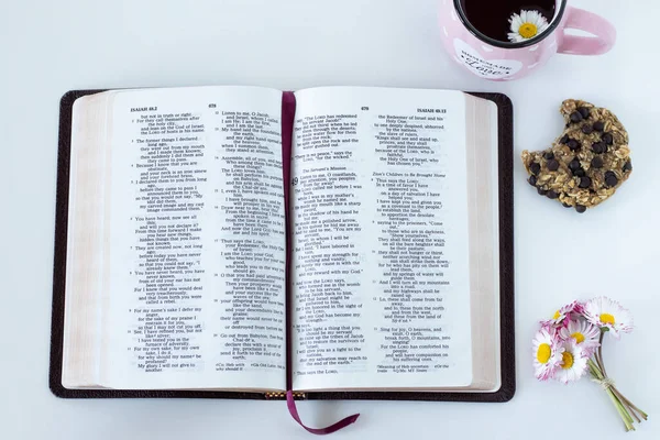 Open Holy Bible Book with a cup of tea, chocolate chip cookie, and spring flowers on a white table. Top view, a close-up. Morning Scripture study and prayer to God Jesus Christ biblical concept.