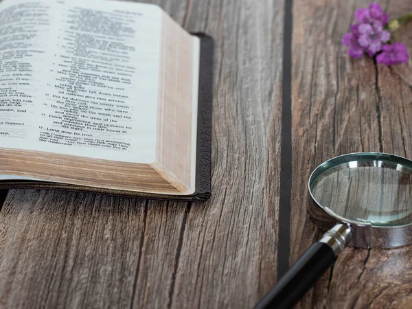 Old open Holy Bible book with a magnifying glass and flower on a wooden table. Search the Scripture, biblical wisdom, and seek the kingdom of God. Matthew 6:33 gospel. A closeup.