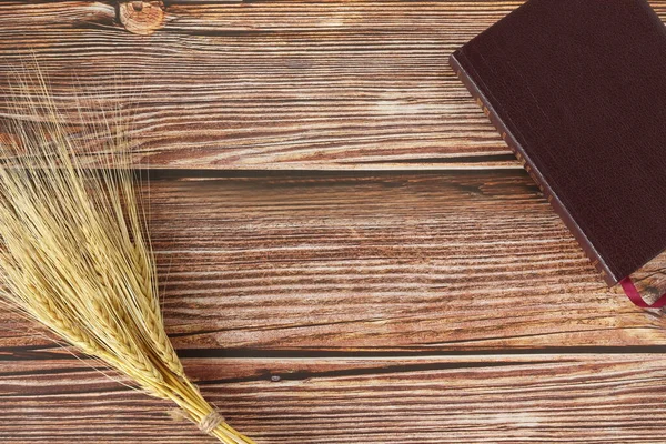 Ripe gold barley sheaf and closed Holy Bible book on wooden table with copy space. Top view. Harvest season, grain offering, Christian thanksgiving, Pentecost festival biblical concept.