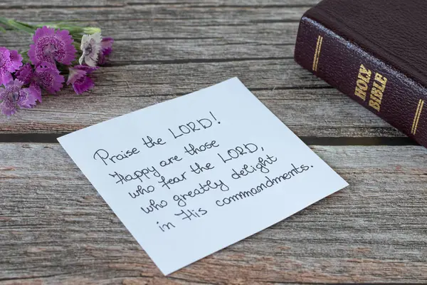 Praise the LORD, handwritten verse on a note, closed Holy Bible Book, and flower on a wooden table. Praise and worship God Jesus Christ biblical concept. Psalm 112. A close-up.
