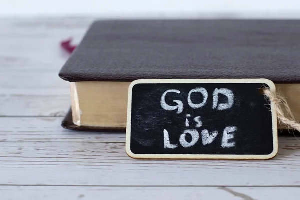 God is love, handwritten verse with chalk on a small blackboard with a closed Holy Bible Book with golden pages in the background. A close-up.