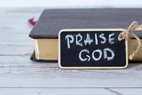 Praise God, a handwritten text on a small blackboard with chalk, and a closed Holy Bible Book with golden pages in the background. Worship the LORD Jesus Christ, Christian biblical concept.
