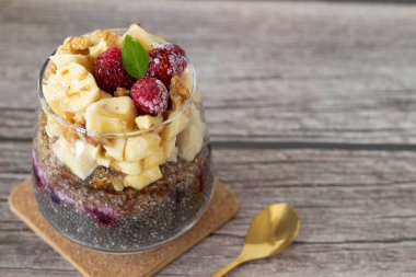 Muz, ahududu ve ahududu dolu bir bardakta vegan chia tohumu pudingi ve fotokopi alanı olan ahşap bir masada altın bir kaşık. - Yakın çekim. Tatlı ve sağlıklı bitki temelli kahvaltı ya da atıştırmalık.