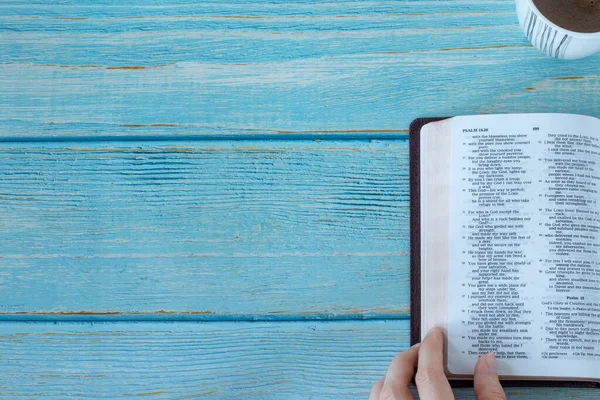 Human hand holding open Holy Bible Book with a cup of coffee on a blue wooden background with copy space. Top table view. Reading and studying the Scripture, Christian biblical concept. A close-up.