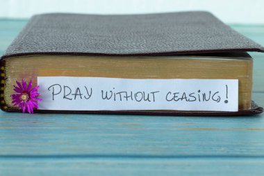 Pray without ceasing, a handwritten text on a note with a purple flower in front of a closed Holy Bible Book with golden pages placed on a blue wooden table with white background. A close-up. clipart