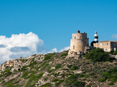 Sardinya 'daki Calamosca Dağı boyunca uzanan ebedi yeşil ağacın yanındaki beyaz deniz feneri kulesi güneşli bir günde mavi gökyüzü ve tüylü beyaz bulutlarla kaplı İtalya' nın Calamosca kenti.