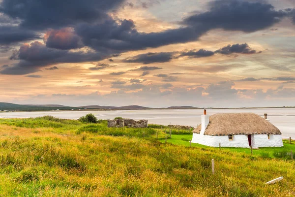 Chalet blanc traditionnellement construit avec toit de chaume, à côté de la baie turquoise, avec un ciel sombre nuageux orageux au-dessus. Île de North Uist, Écosse — Photo