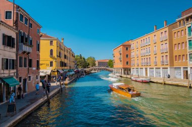VENICE, İtalya - 27 Ağustos 2021: Venedik Kanalı 'nın yanındaki kaldırımda taksi botu ve diğer teknelerle yürüyen turistleri görmek.