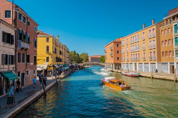 VENICE, İtalya - 27 Ağustos 2021: Venedik Kanalı 'nın yanındaki kaldırımda taksi botu ve diğer teknelerle yürüyen turistleri görmek.