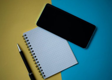 Ofis işyeri konsepti çok az. Kurşun kalemli boş bir defter, sarı ve mavi arka planda cep telefonu. Kopyalama alanı olan üst görünüm, düz yatay
