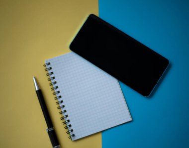 Ofis işyeri konsepti çok az. Kurşun kalemli boş bir defter, sarı ve mavi arka planda cep telefonu. Kopyalama alanı olan üst görünüm, düz yatay