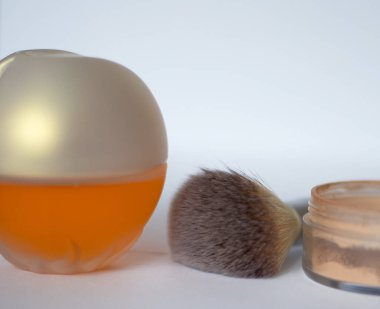 Sarı parfüm şişesi, yüz pudrası ve beyaz arka planda makyaj fırçası.