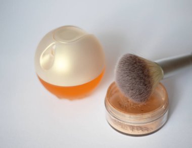 Sarı parfüm şişesi, yüz pudrası ve beyaz arka planda makyaj fırçası.