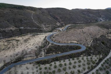 Dağlık bir araziyi geçen yollar, zeytin ve badem ağaçları yetiştiriliyor, çalılar var, gökyüzü açık.