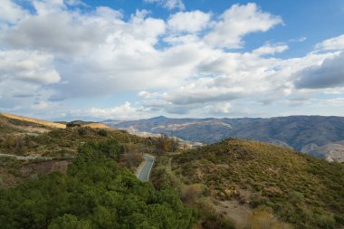 İspanya 'nın güney Endülüs bölgesindeki dağlık bölgede bitki örtüsü var, vadi var, dağın yanından geçen bir yol var, gökyüzünde bulutlar var.