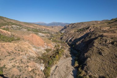 mountainous landscape in the south of Spain, there are trees and bushes, a road passes by the side of the mountain, the sky is clear