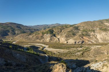 mountainous landscape in the south of Spain, there are trees and bushes, a road passes by the side of the mountain, the sky is clear
