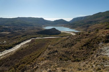 mountainous landscape in southern Spain, there are trees and bushes, there is a water reservoir, the sky is clear