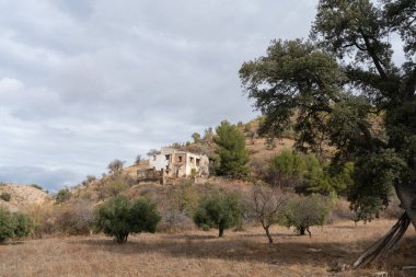 İspanya 'nın güneyindeki dağlık arazi, ağaçlar ve çalılar var, bir kır evi var, gökyüzü bulutlu.