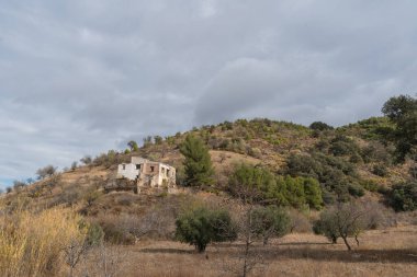 İspanya 'nın güneyindeki dağlık arazi, ağaçlar ve çalılar var, bir kır evi var, gökyüzü bulutlu.