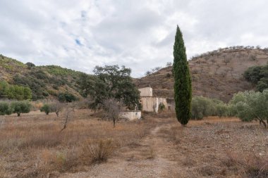 İspanya 'nın güneyinde dağlık bir arazi, ağaçlar ve çalılar var, bir kır evi var, toprak yollar var, gökyüzü bulutlu.