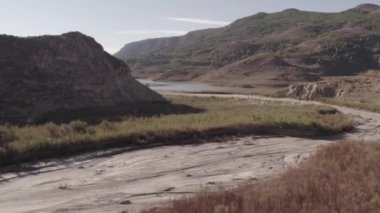 Çalı ve ağaçlardan oluşan bir ormanın üzerinde uçan, dağlık bir alan, bir su deposu var, gökyüzünde çok az bulut var