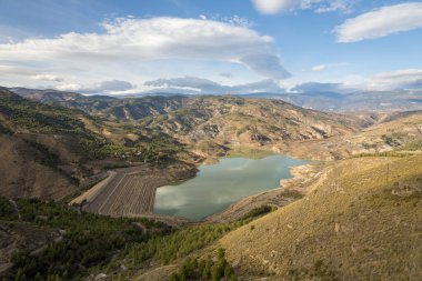 İspanya 'nın güneyinde Beninar rezervuarı var. Ağaçlı ve çalılı dağlar var. Gökyüzünde bulutlar var.