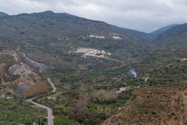 Güney Endülüs 'ün dağlık bölgelerinde ağaçlar ve çalılar var, vadide birkaç çiftlik evi var, yamaçta birkaç köy var, gökyüzünde bulutlar var.