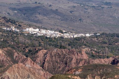Dağın kenarındaki bir kasaba, evlerin cepheleri beyaza boyanmış, ağaçlar ve çalılar var, sarp alanlar var.