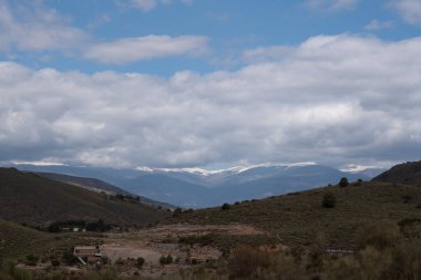 İspanya 'nın güneyindeki dağlık arazi, dağlarda kar, çalılar ve ağaçlar, birkaç çiftlik evi, gökyüzünde bulutlar var.
