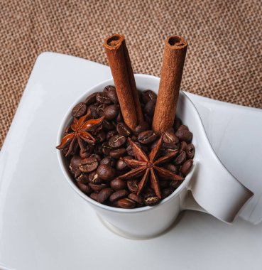 Masanın üzerinde beyaz bir fincan tabağı. Bardakta kahve çekirdekleri, iki tarçın çubuğu ve iki yıldız anason var. Yukarıdan bak. Boşluğu kopyala..