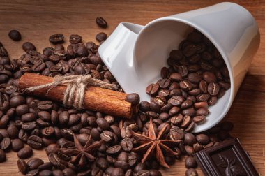 Tahta bir masanın üzerinde kahve çekirdekleri olan beyaz bir fincan var. Fincanın yanında da kahve çekirdekleri, tarçın çubukları, anason yıldızları ve bir parça çikolata var. Boşluğu kopyala..