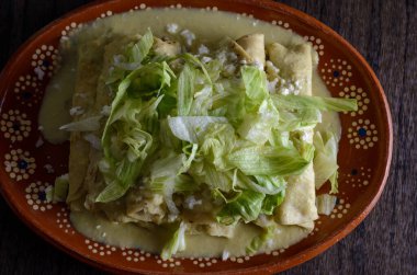Green Enchiladas with lettuce served in a clay dish on a wooden table.