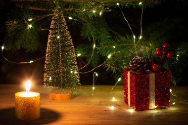 Kırmızı bir Noel kutusu, yanan bir mum ve arka planda Noel ışıkları olan doğal bir köknar ağacı olan küçük bir Noel ağacı.