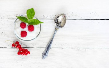 Sütlü yoğurt, olgun ahududu, yeşil yaprak ve kırmızı frenk üzümlü beyaz ahşap masa üzerinde bir bardak.