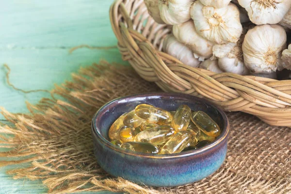 Transparent golden garlic oil capsules in ceramic bowl and fresh garlic in basket on sackcloth and on green old wooden background.