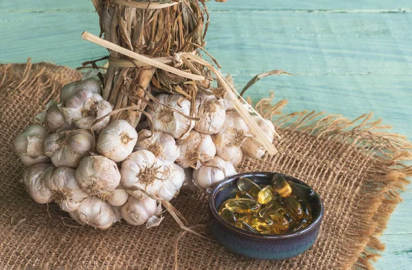 Transparent golden garlic oil capsules in ceramic bowl, garlic bulbs on sackcloth and on green old wooden background.