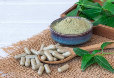 Andrographis, tahta kaşıkta paniklemiş bitkisel kapsüller, yeşil kasede, kahverengi çuvalda ve beyaz ahşap arka planda paniklemiş toz..